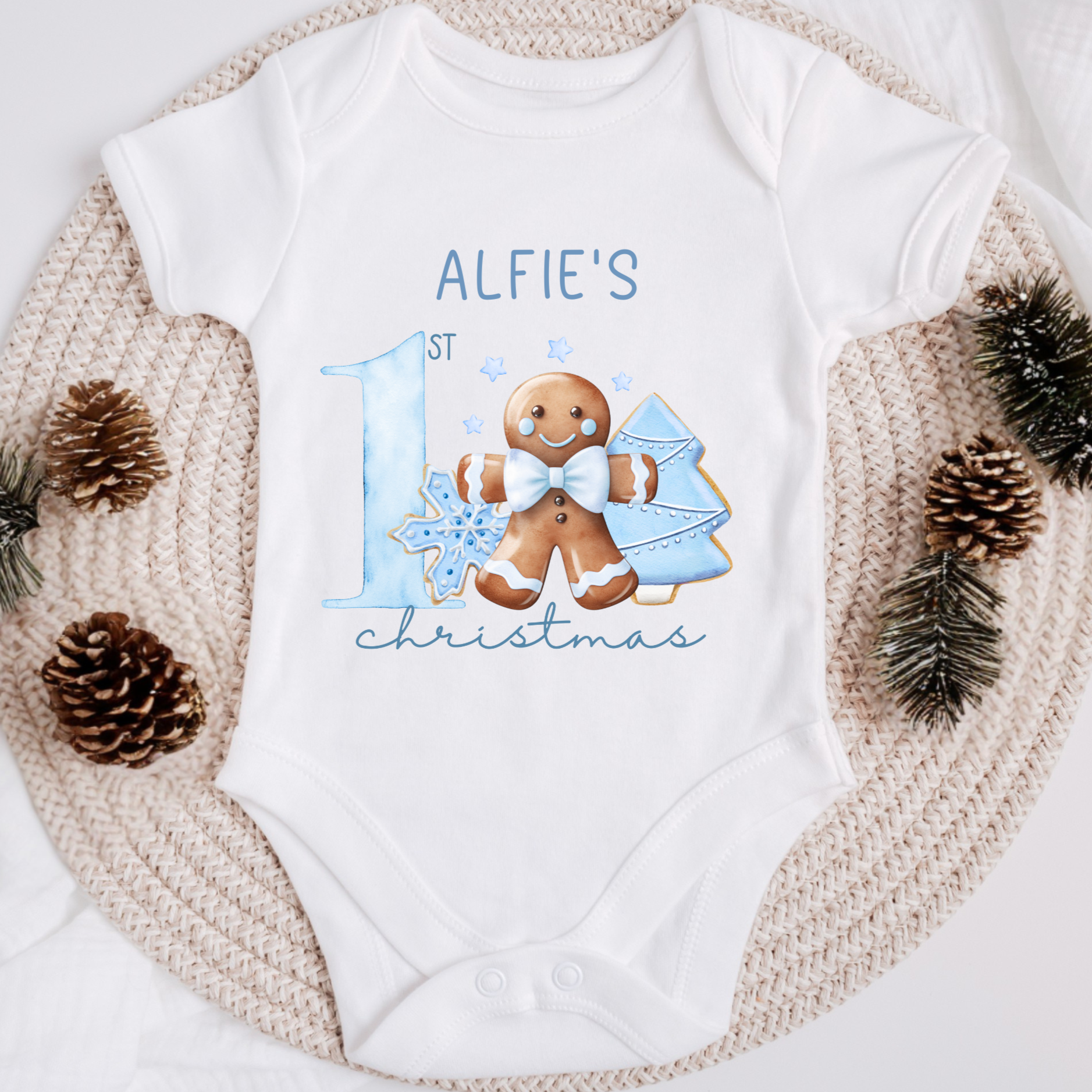 White baby onesie with gingerbread design and text on a textured surface with pinecones and branches.