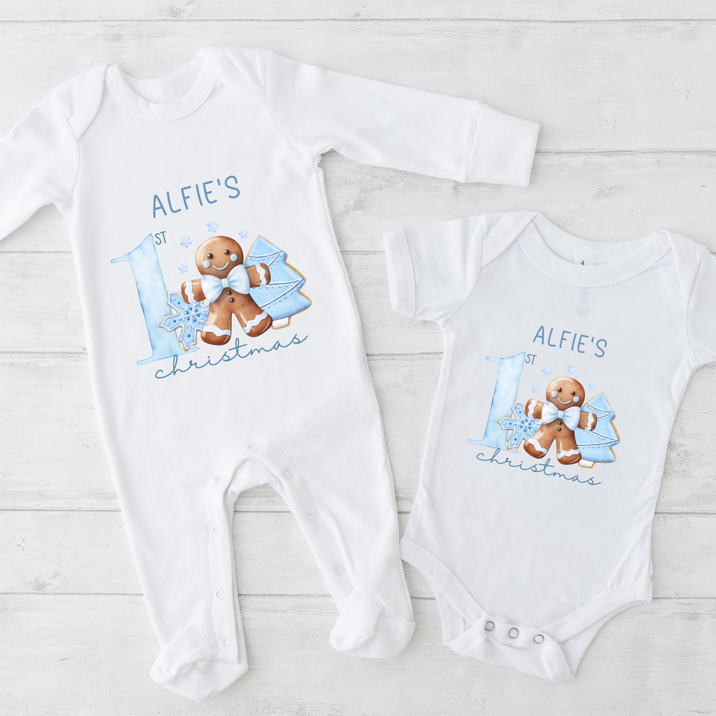 Two baby onesies with a gingerbread man design and text on a light wooden surface.